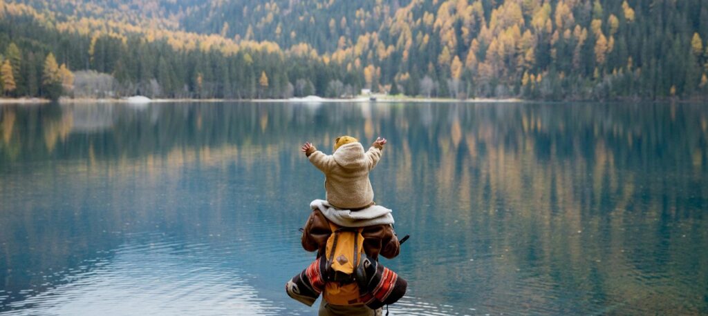 Padre cargando a su hijo pequeño sobre los hombros frente a un lago rodeado de montañas